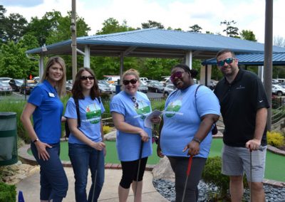 A group of 5 people smiling at the camera in front of a putting green. On the left, a woman in a royal blue collared shirt with a name tag. Next to her, a woman with long brown hair and sunglasses wearing a light blue event shirt with a golf graphic on the front is holding a mini-golf club. Next to her, a woman with a blonde pony tail and sunglasses, and a matching event shirt holds a mini-golf club. Next to her, a black woman with her hair pulled back into a bun wears sunglasses, a matching event shirt and also holds a mini-golf club. On the far right, a man with a beard and mustache and sunglasses wears a black shirt and holds a mini-golf club.