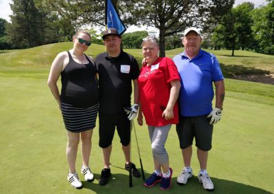 Two couples smile for the camera on the golf course. One woman is wearing a red Associa Cares shirt.