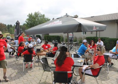 People eating lunch and chatting on a patio with tables and umbrellas in front of a golf course. Many of the people are wearing red Associa Cares shirts.