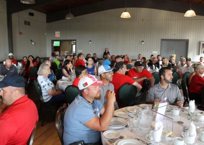 A group of people, some wearing red Associa cares shirts, watch a speaker from their dinner tables inside a large building at the golf course.
