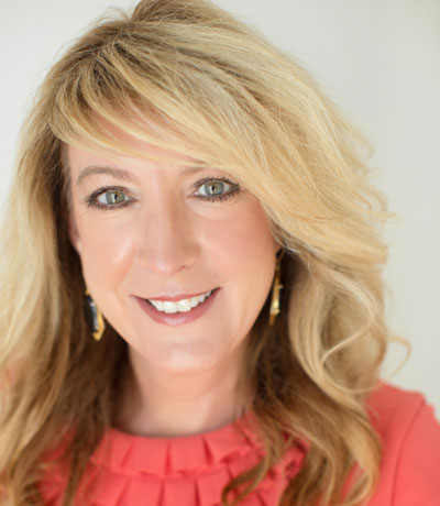 Headshot of Lisa Locke, a woman with long, layered, blonde hair, gold earrings, green eyes, and a coral colored blouse.