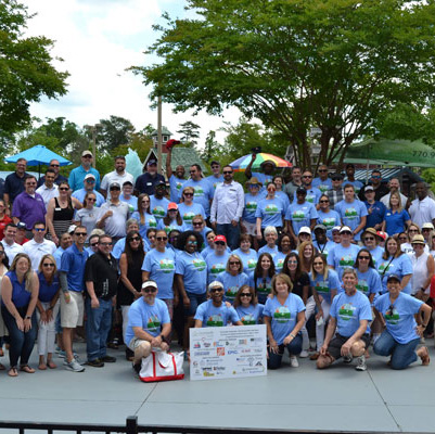 A group photo of 70+ people wearing matching blue shirts at a volunteer event. The person in front is holding a sign with a list of sponsors.