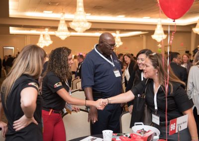A woman with red pants and a black shirt standing behind her conference table shakes hands with another woman wearing a black shirt. The room is full of people chatting with each other and smiling. A red balloon and some swag adorns the conference table.