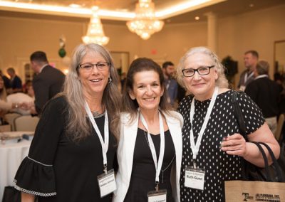 Three women smile for the camera at a conference. People behind them are mingling and smiling. The names from left to right (based on their name tags) are Debbie Johnson, a woman with long grey hair and a black blouse, Ms. Campbell, a woman with long, tied-back hair, a black shirt and a white suit jacket, and Ruth Gunn, a woman with curly gray hair, tied-back, and a black and white polka-dot blouse.