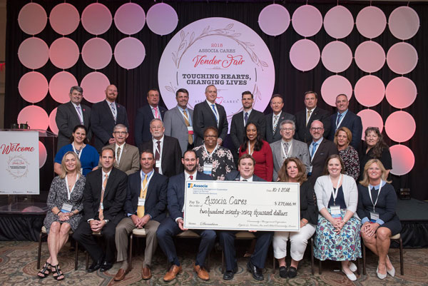 A group of women and men dressed in business attire pose behind an oversized check from Associa that says "Pay to the order of Associa Cares. $277,000. October 1, 2018." The group stands in front of a sign that says "2018 Associa Cares Vendor Fair. Touching Hearts, changing lives"