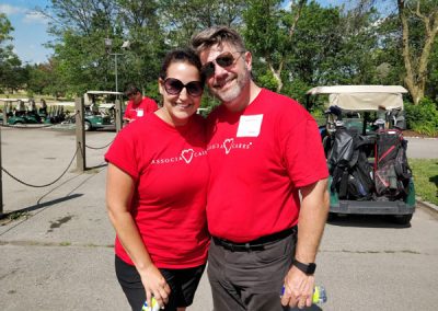 A man and a woman wearing red Associa Cares shirts smile at the camera in front of a row of golf carts.