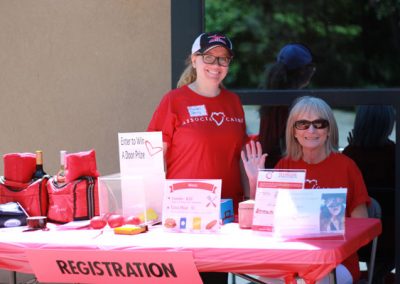 Two women wearing red Associa Cares shirts sit behind a table with a red table cloth that says "Registration" and "Enter to win a door prize."