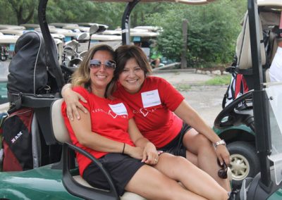 A woman puts her arm around another woman in a golf cart and they smile at the camera. They are wearing red Associa Cares shirts and name tags.