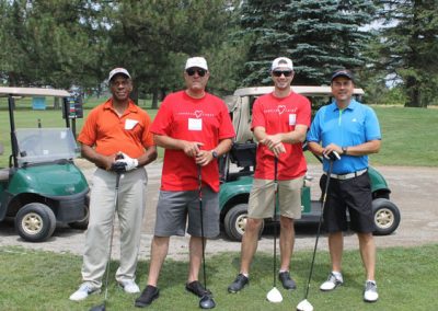 Four men stand in front of golf carts holding golf clubs. Two of the men are wearing Associa Cares shirts.