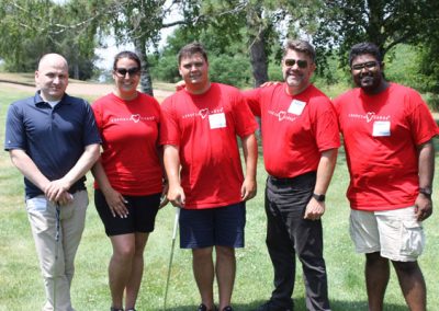 A group of 4 men and 1 woman smiling on a golf course. The woman and 3 men wear red Associa Cares shirts.