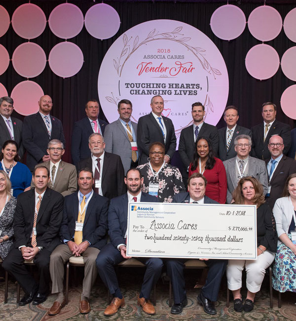 A group of women and men dressed in business attire pose behind an oversized check from Associa that says "Pay to the order of Associa Cares. $277,000. October 1, 2018." The group stands in front of a sign that says "2018 Associa Cares Vendor Fair. Touching Hearts, changing lives"